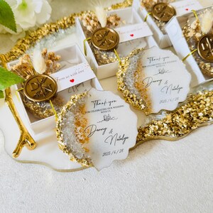 May include: Wedding favors with gold glitter and a white background. The favors are small, round, and have a clear resin top with gold flakes. The text on the favors reads "Thank you for celebrating our wedding with us, Danny & Natalya, 2025/6/28".