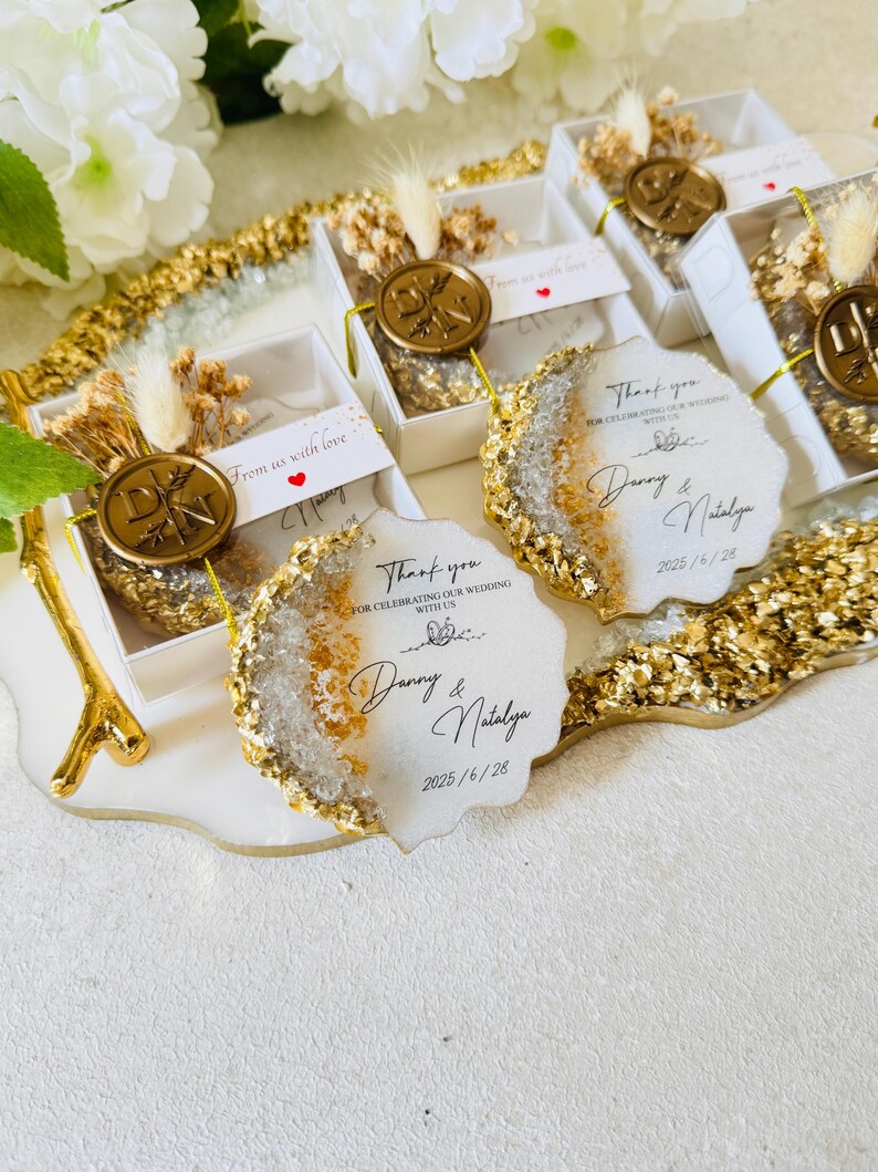 May include: Wedding favors with gold glitter and a white background. The favors are small, round, and have a clear resin top with gold flakes. The text on the favors reads "Thank you for celebrating our wedding with us, Danny & Natalya, 2025/6/28".