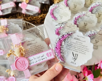 May include: A set of small, white, decorative favors with pink and silver accents. The favors are shaped like a crescent moon and feature a pink and silver glitter design. The favors are packaged in clear boxes with pink ribbons and include a small, white card with Arabic text. The text on the card reads "Allahumma Inbit-ha Nabatan Hasanaw Wa-j'al-ha Qurat-a 'Aynin Li-Waliday-ha" and "Ayla".