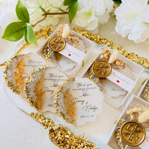 May include: Wedding favors in small clear boxes with gold accents. Each box contains a small white and gold resin coaster with a gold wax seal and dried flowers. The boxes have a thank you note with the couple's names and wedding date.