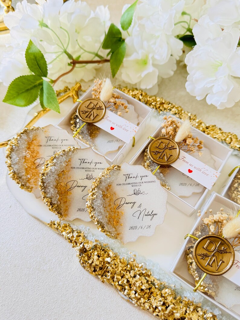 May include: Wedding favors in small clear boxes with gold accents. Each box contains a small white and gold resin coaster with a gold wax seal and dried flowers. The boxes have a thank you note with the couple's names and wedding date.
