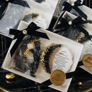 May include: Gift boxes with black bows and gold accents. Each box contains a black and gold decorative item, a prayer bead necklace, and a small card with Arabic text. A gold seal with the text "Hajj Mabroor" is also included.