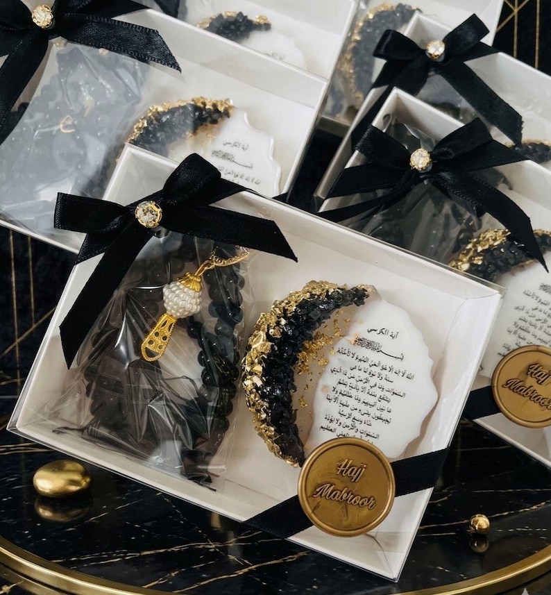 May include: Gift boxes with black bows and gold accents. Each box contains a black and gold decorative item, a prayer bead necklace, and a small card with Arabic text. A gold seal with the text "Hajj Mabroor" is also included.