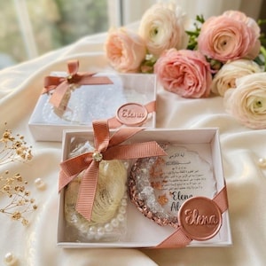 May include: Two white gift boxes tied with peach ribbons and embellished with a gold gem. One box contains a pearl necklace and a gold-colored item. The other box contains a white item. Each box has a circular seal that says "Elena".