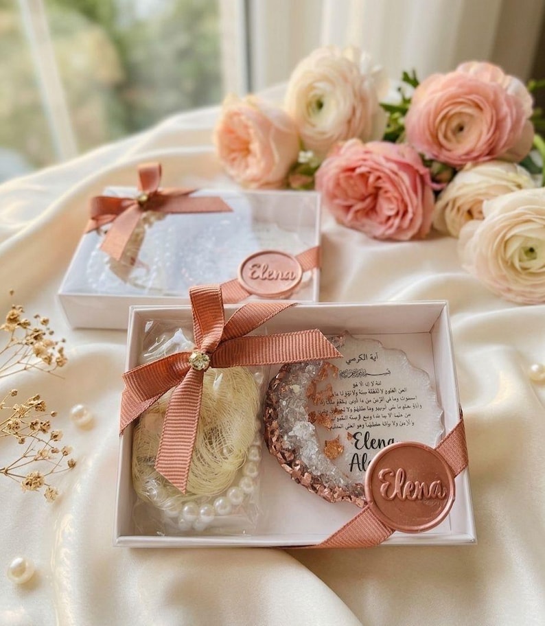 May include: Two white gift boxes tied with peach ribbons and embellished with a gold gem. One box contains a pearl necklace and a gold-colored item. The other box contains a white item. Each box has a circular seal that says "Elena".