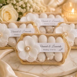Pode incluir: Elegantes lembrancinhas de casamento em bandejas douradas, cada uma contendo doces brancos. As bandejas são adornadas com detalhes florais brancos, pérolas e um rótulo que diz "Hannah & Leonardo Wedding 07.07.26". Uma vela e flores estão no fundo.