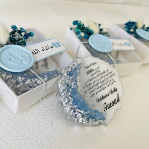 May include: A white and blue baby shower favor box with a blue and silver resin ornament. The ornament has a white background with Arabic text and the words "Welcome Baby Tawad".