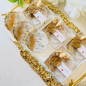 May include: A tray of small white boxes with clear lids, each containing a white card with gold lettering and a small dried flower arrangement. The boxes are decorated with gold glitter and the cards say "Thank you for celebrating our wedding with us" and "From us with love".