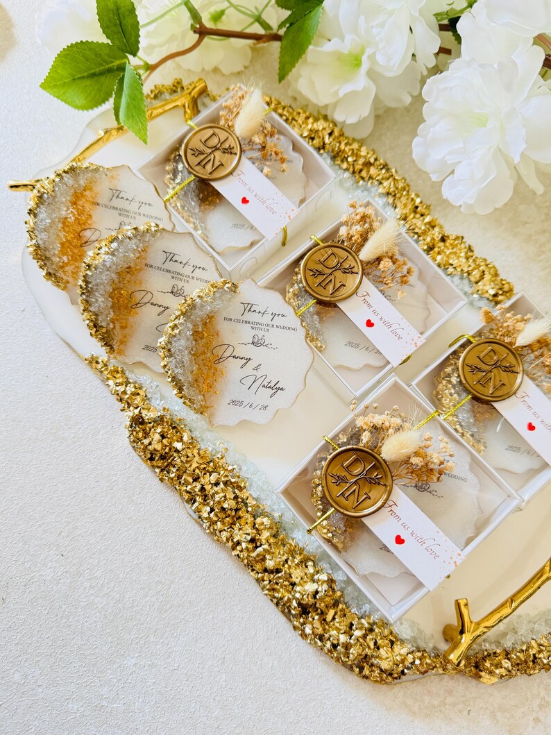 May include: A tray of small white boxes with clear lids, each containing a white card with gold lettering and a small dried flower arrangement. The boxes are decorated with gold glitter and the cards say "Thank you for celebrating our wedding with us" and "From us with love".