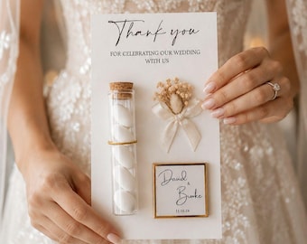 May include: A wedding favor card held by a person. The card reads "Thank you" and includes a glass tube with white candies, a small floral arrangement, and a framed name card with the date.