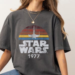May include: A dark gray t-shirt with a vintage-style Star Wars graphic. The graphic features a spaceship flying through a sunset with the text "Star Wars 1977".