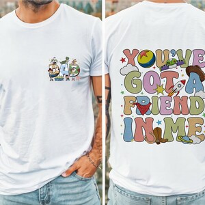 May include: White t-shirt with a colorful graphic design. The front features the word "DAD" with cartoon characters. The back displays the phrase "YOU'VE GOT A FRIEND IN ME" with various illustrations, including a rocket and cowboy boots.