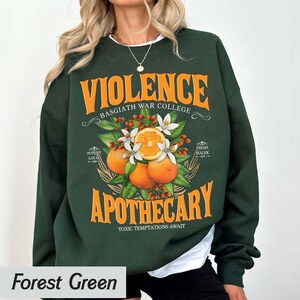 Puede incluir: Sudadera verde bosque con la palabra "VIOLENCE" en naranja, encima de "BASGIATH WAR COLLEGE". Debajo, "APOTHECARY" est&aacute; impreso en naranja, con una ilustraci&oacute;n de naranjas, flores y hojas. El texto "TOXIC TEMPTATIONS AWAIT" est&aacute; debajo.