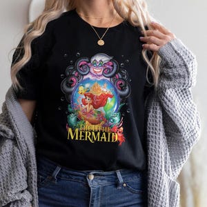 May include: Black t-shirt featuring a colorful graphic from "The Little Mermaid." The design includes Ariel, Ursula, and other characters within a bubble. The words "The Little Mermaid" are printed in yellow and red.