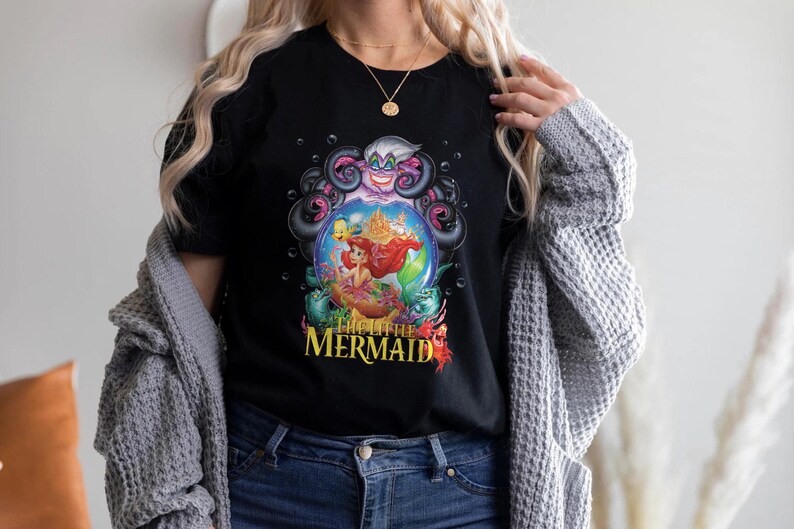 May include: Black t-shirt featuring a colorful graphic from "The Little Mermaid." The design includes Ariel, Ursula, and other characters within a bubble. The words "The Little Mermaid" are printed in yellow and red.