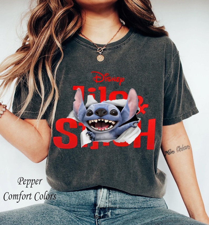 Puede incluir: Camiseta gris oscuro con el personaje de Disney Stitch rompiendo la camiseta, con las palabras "Lilo & Stitch" en rojo. La camiseta tambi&eacute;n tiene las palabras "Pepper Comfort Colors" impresas.