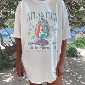 May include: White t-shirt featuring a graphic of the Little Mermaid with the text "Atlantica" and "Little Mermaid". The design includes a castle, seashells, and the year "1989". The shirt has short sleeves and a relaxed fit.