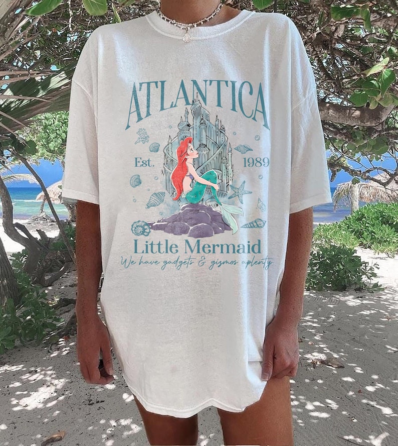 May include: White t-shirt featuring a graphic of the Little Mermaid with the text "Atlantica" and "Little Mermaid". The design includes a castle, seashells, and the year "1989". The shirt has short sleeves and a relaxed fit.