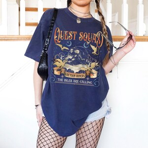 Peut inclure: T-shirt bleu marine surdimensionn&eacute; avec l'inscription "Quest Squad" au-dessus d'une illustration de montagne avec des oiseaux et le texte "Aretian Riders, The Isles Are Calling". Le t-shirt est port&eacute; avec un short en jean et des bas r&eacute;sille.