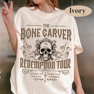 May include: Ivory-colored t-shirt featuring a graphic design with the text "THE BONE CARVER REDEMPTION TOUR." The design includes a skull with crossed swords and decorative scrollwork. The shirt also lists "FEATURING THE DEATH-GOD" and "SPECIAL GUEST THE WEAVER."