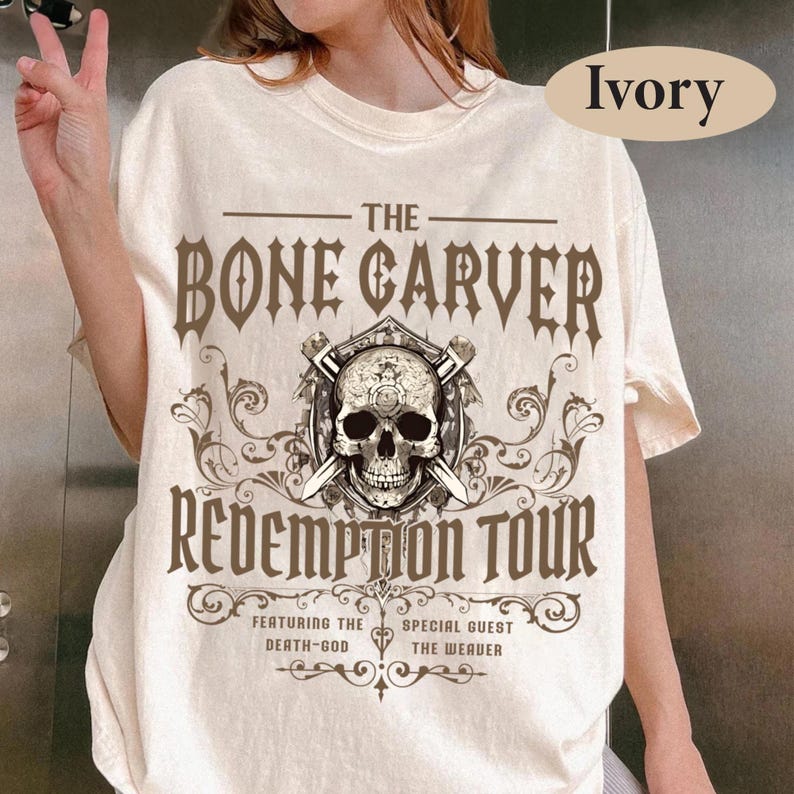 May include: Ivory-colored t-shirt featuring a graphic design with the text "THE BONE CARVER REDEMPTION TOUR." The design includes a skull with crossed swords and decorative scrollwork. The shirt also lists "FEATURING THE DEATH-GOD" and "SPECIAL GUEST THE WEAVER."
