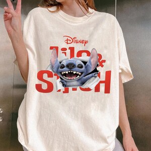 Puede incluir: Camiseta color marfil con el logo de Disney y las palabras "Lilo & Stitch" en rojo. El personaje azul Stitch rompe el texto. La camiseta tiene un corte holgado. La palabra "IVORY" est&aacute; impresa en la parte inferior.