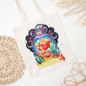 May include: Cream-colored tote bag featuring a colorful graphic from Disney's The Little Mermaid. The design includes Ariel, Ursula, and other characters. The text "THE LITTLE MERMAID" is printed in yellow. The bag has two cream-colored handles.