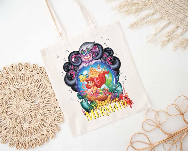 May include: Cream-colored tote bag featuring a colorful graphic from Disney's The Little Mermaid. The design includes Ariel, Ursula, and other characters. The text "THE LITTLE MERMAID" is printed in yellow. The bag has two cream-colored handles.