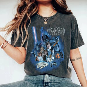 May include: A dark gray t-shirt with a vintage-style Star Wars graphic featuring characters like Darth Vader, Luke Skywalker, Chewbacca, and Princess Leia. The text "STAR WARS" is printed in white above the graphic.