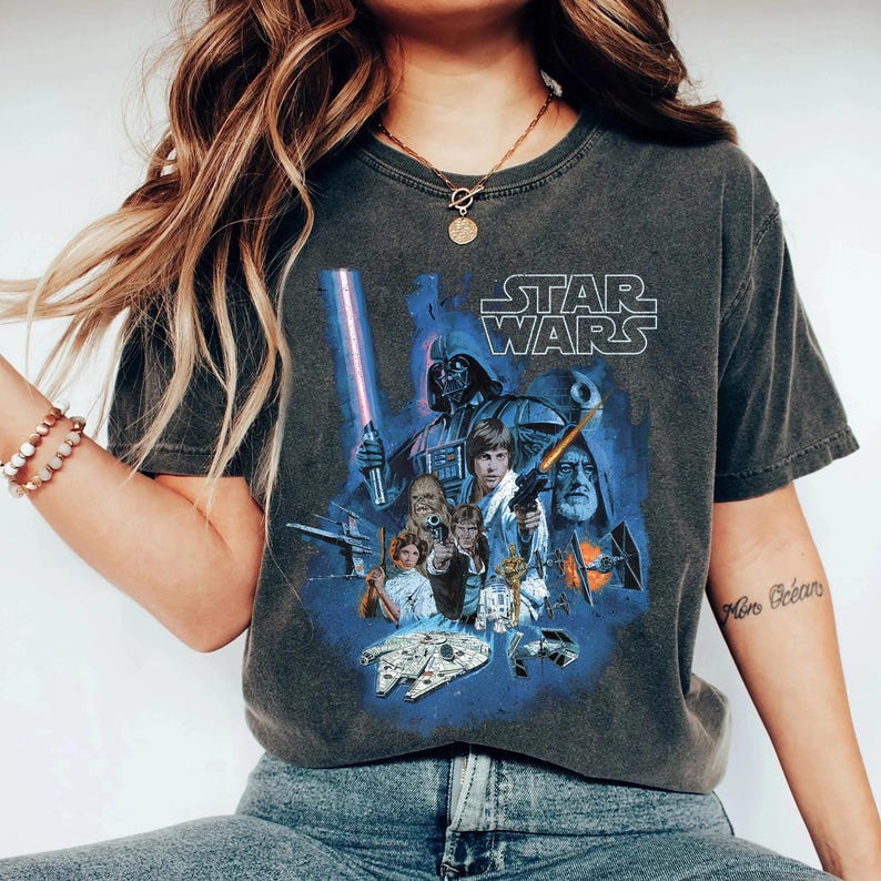 May include: A dark gray t-shirt with a vintage-style Star Wars graphic featuring characters like Darth Vader, Luke Skywalker, Chewbacca, and Princess Leia. The text "STAR WARS" is printed in white above the graphic.