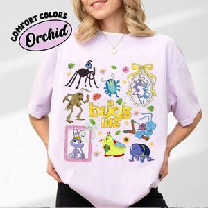 May include: Orchid Comfort Colors t-shirt featuring a colorful graphic of characters from "A Bug's Life." The design includes various cartoon insects and the text "a bug's life" in yellow. The shirt is a light purple color.