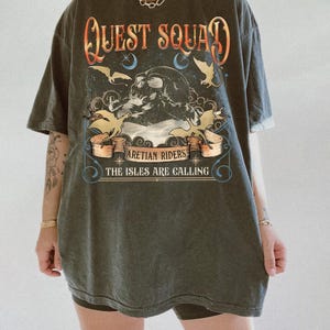 Peut inclure: T-shirt gris fonc&eacute; avec un motif graphique. Le motif comprend le texte "QUEST SQUAD" en orange et or, un cr&acirc;ne, des chauves-souris et le texte "ARETIAN RIDERS THE ISLES ARE CALLING". Le t-shirt a un aspect vintage et us&eacute;.