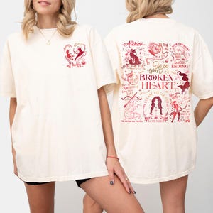 May include: Cream-coloured t-shirt with red graphic designs. The front features a heart-shaped design with the word "Archer". The back displays illustrations and text, including "Once upon a Broken Heart".