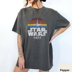 May include: A dark gray t-shirt with a vintage-style Star Wars graphic. The graphic features a silhouette of an X-Wing fighter flying over a sunset with the text "Star Wars 1977".