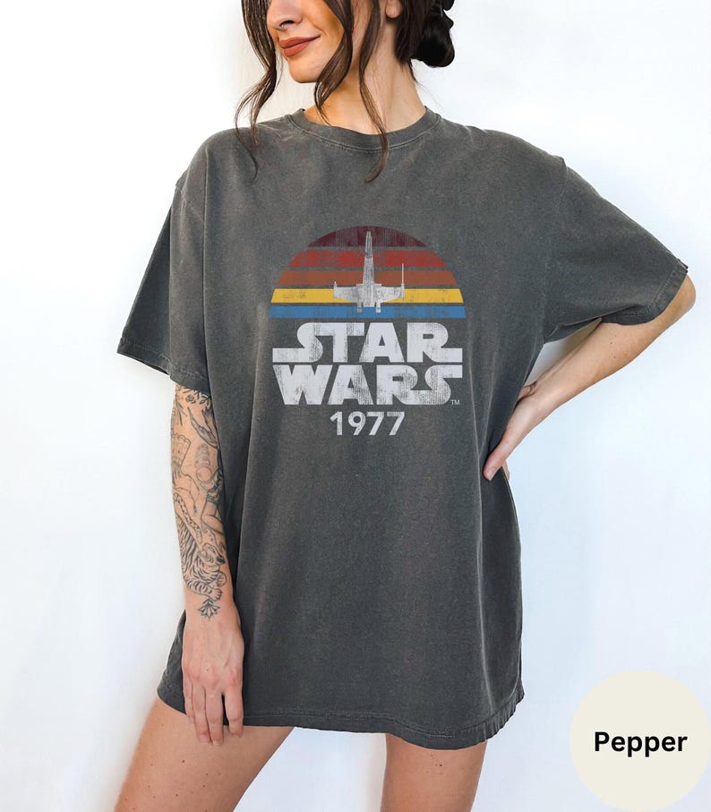 May include: A dark gray t-shirt with a vintage-style Star Wars graphic. The graphic features a silhouette of an X-Wing fighter flying over a sunset with the text "Star Wars 1977".
