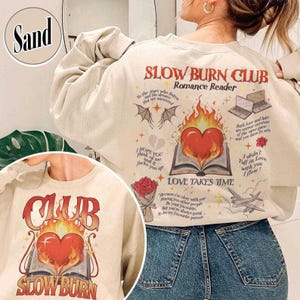 May include: Sand-colored sweatshirt with the text "SLOW BURN CLUB Romance Reader" in red and orange. The design includes a heart on fire, an open book, and various romantic-themed illustrations. The sweatshirt also has the text "LOVE TAKES TIME".
