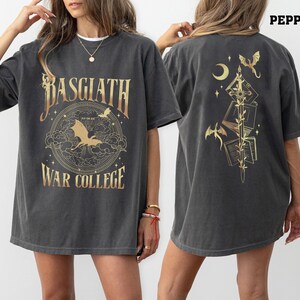 Könnte beinhalten: Dunkelgraues T-Shirt mit goldfarbenem Schriftzug und Grafiken. Die Vorderseite zeigt den Text "BASGIATH WAR COLLEGE" mit einem Drachen und einem kreisförmigen Design. Die Rückseite hat ein Schwertdesign mit Büchern, Drachen und einem Halbmond.