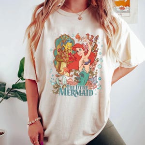 May include: Cream-colored t-shirt featuring a colorful graphic of Ariel from The Little Mermaid. The design includes Ariel with red hair and a teal tail, along with Flounder and the text "The Little Mermaid". The shirt has short sleeves.