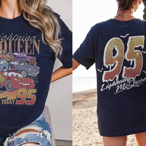 K&ouml;nnte beinhalten: Marineblaues T-Shirt mit einem Aufdruck von Lightning McQueen und anderen Cars-Charakteren, dem Text "Trick or Treat" und "95". Auf der R&uuml;ckseite des Shirts ist eine gro&szlig;e "95" und der Text "Lightning McQueen" zu sehen.