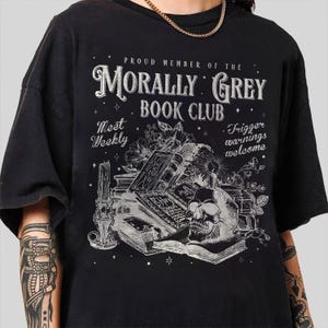 May include: Black t-shirt with a graphic design featuring a skull, books, and floral elements. The text reads "PROUD MEMBER OF THE MORALLY GREY BOOK CLUB" with additional text "Meet Weekly" and "Trigger warnings welcome."