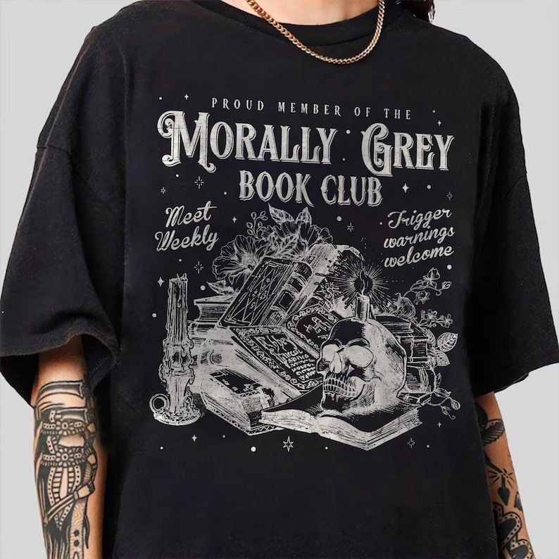 May include: Black t-shirt with a graphic design featuring a skull, books, and floral elements. The text reads "PROUD MEMBER OF THE MORALLY GREY BOOK CLUB" with additional text "Meet Weekly" and "Trigger warnings welcome."