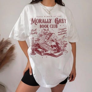 May include: White t-shirt with a maroon graphic featuring a skull, books, and floral elements. The text reads "Proud Member of the Morally Grey Book Club" with additional phrases "Meet Weekly" and "Trigger warnings welcome."