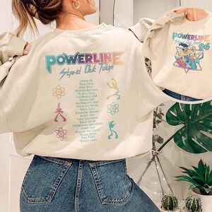 K&ouml;nnte beinhalten: Cremefarbenes Sweatshirt mit einem Powerline Stand Out Tour-Aufdruck auf dem R&uuml;cken und einem kleineren Powerline '96-Aufdruck auf der Vorderseite. Das Design zeigt farbenfrohe Schrift und Illustrationen, die eine Retro-Konzert-Atmosph&auml;re erzeugen.