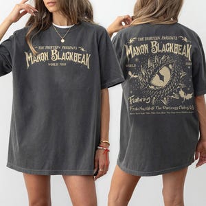 May include: Dark grey t-shirt with the text "THE THIRTEEN PRESENTS MANON BLACKBEAK WORLD TOUR" on the front. The back of the shirt features a graphic design with the text "Featuring From Now Until The Darkness Claims Us".