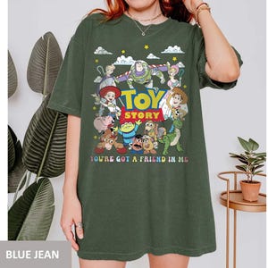 May include: Dark green t-shirt featuring a colorful graphic of characters from the animated film "Toy Story." The design includes the film's title and the phrase "You've Got a Friend in Me." The shirt has short sleeves and a relaxed fit.
