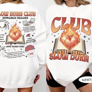 May include: White sweatshirt with a graphic design featuring the text "SLOW BURN CLUB" and "ROMANCE READER." The design includes a heart engulfed in flames above an open book. Additional text and illustrations are on the back.