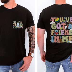 May include: Black t-shirt with a colorful graphic on the front that says "DAD" with cartoon characters. The back of the shirt has the phrase "YOU'VE GOT A FRIEND IN ME" in a retro font with cartoon images. The shirt is short-sleeved.