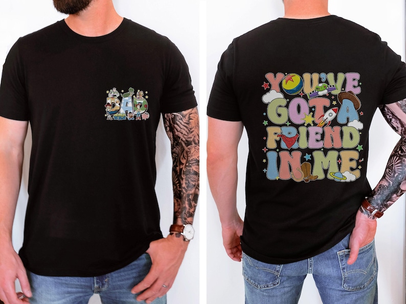 May include: Black t-shirt with a colorful graphic on the front that says "DAD" with cartoon characters. The back of the shirt has the phrase "YOU'VE GOT A FRIEND IN ME" in a retro font with cartoon images. The shirt is short-sleeved.
