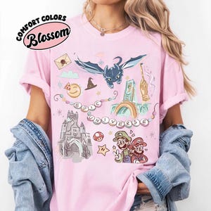 May include: Pale pink Comfort Colors t-shirt with a graphic design featuring a dragon, castle, and video game characters. The text "EPIC UNIVERSE" is displayed across the front. The shirt also has the text "Blossom" in a circular design.