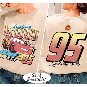 May include: Sand-colored sweatshirt featuring Lightning McQueen from the Cars movie. The front graphic shows McQueen with text "Lightning McQueen" and "FAST 95". The back displays a large "95" and "Lightning Sally" text.